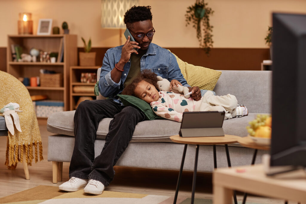 Wide shot of sick child lying on couch and hugging toy looking at tablet while her concerned father talking on phone with pediatrician